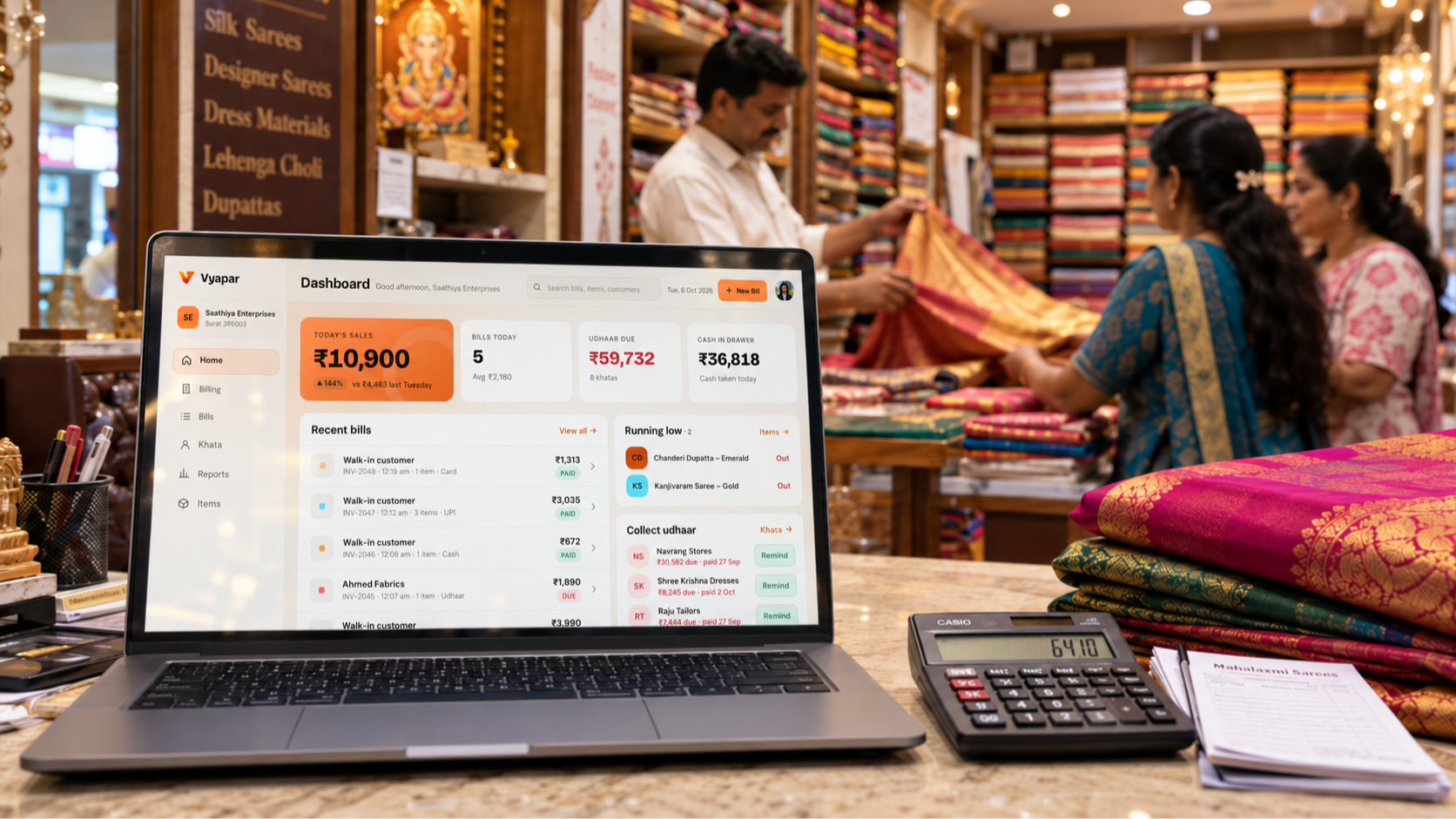 Vyapar billing software running on a laptop at an Indian saree shop counter, with a calculator, folded sarees, and customers browsing fabrics in the background.